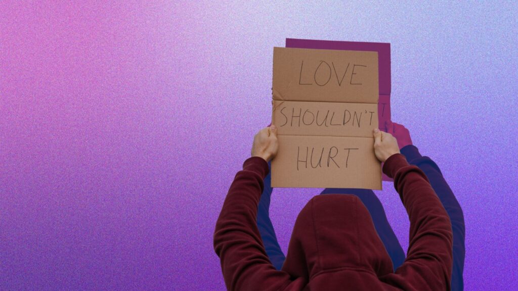 Eine Person mit übergezogener Kapuze steht mit dem Rücken zur Betrachterin. Die Person hält das Schild "Love shouldn't hurt" in die Höhe.